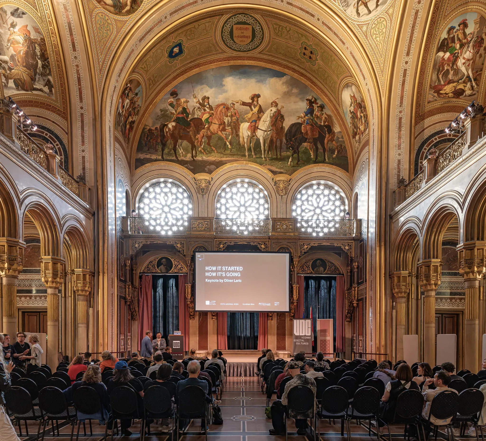 Historischer Saal mit Deckengemälde, Publikum vor einer Leinwand.